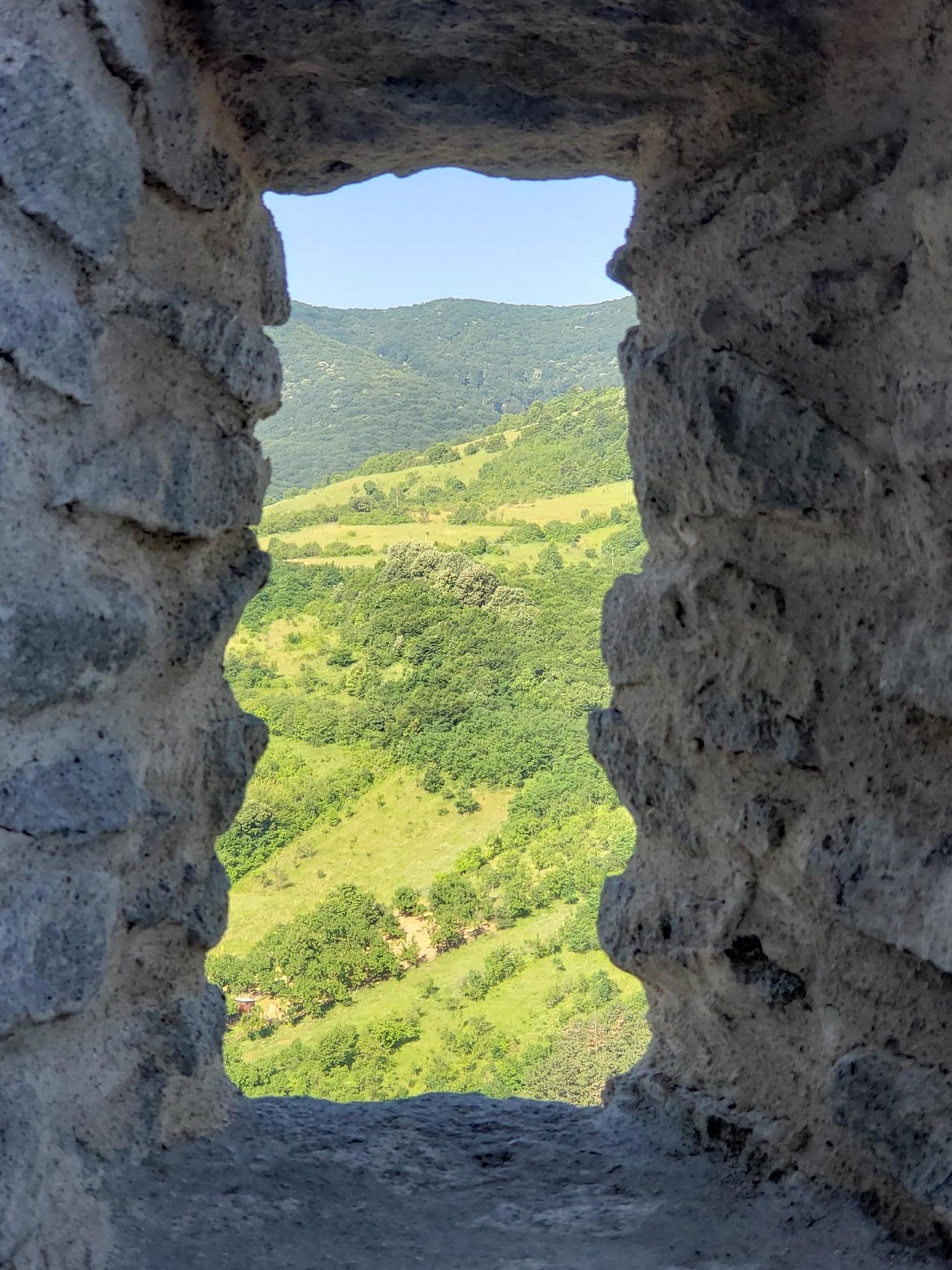 A rectangular opening in an old and thick stone wall reveals a view of green mountains and a blue sky.