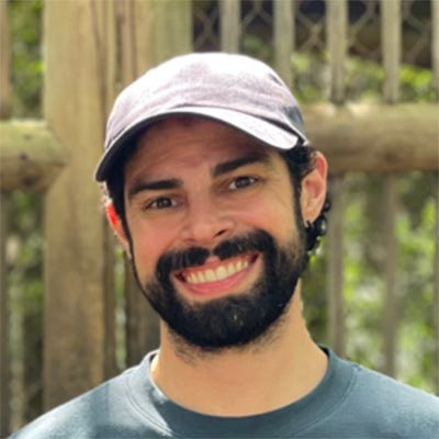 Homem sorridente, com barba, usando um boné claro e uma camiseta cinza-azulada de mangas curtas. Ele está em um ambiente ao ar livre, com vegetação ao fundo. Homem sorridente, com barba, usando um boné claro e uma camiseta cinza-azulada de mangas curtas. Ele está em um ambiente ao ar livre, com vegetação ao fundo.