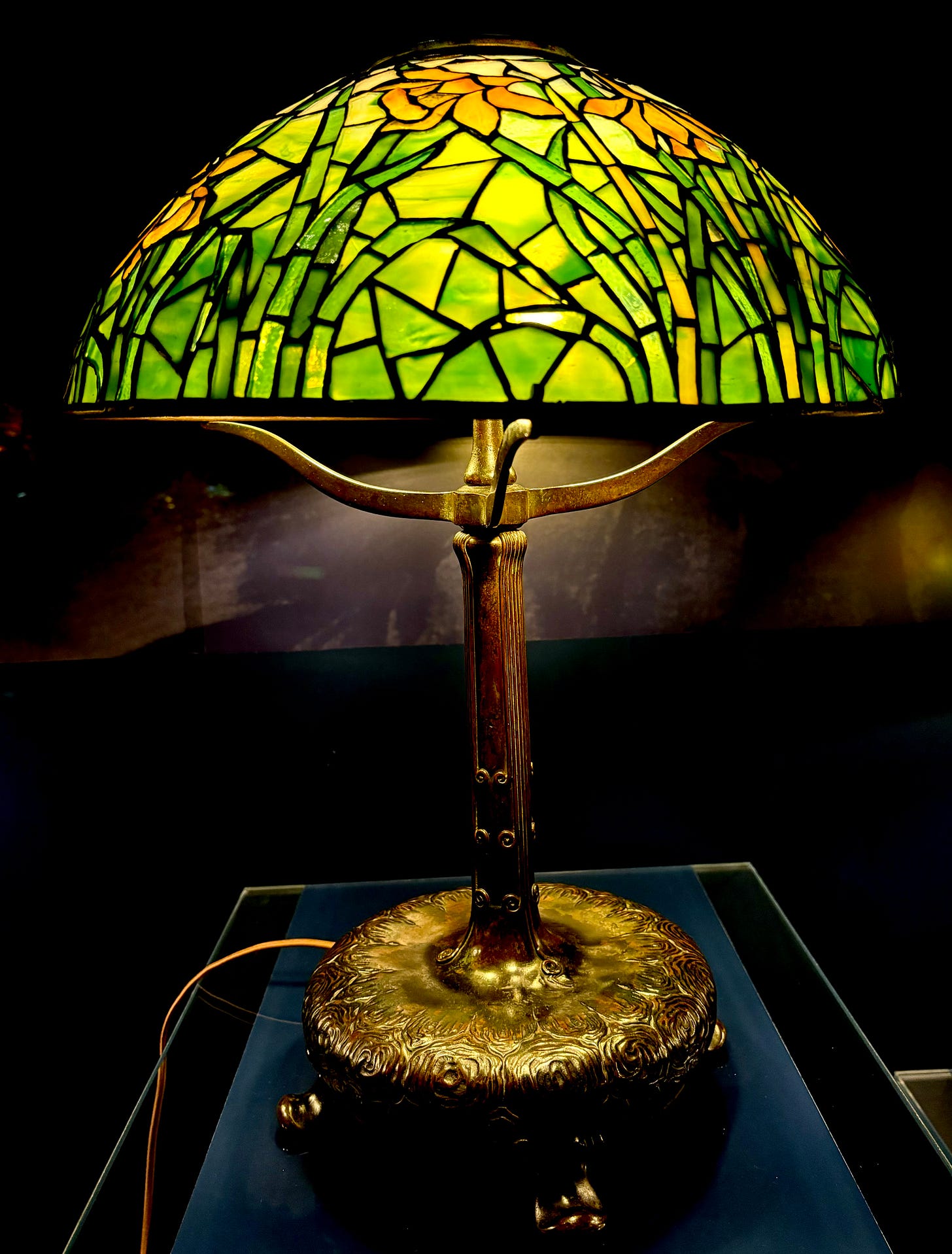 A metal base with convex arms supporting a glass base. It is green with distinct leaves of glass and yellow daffodils.