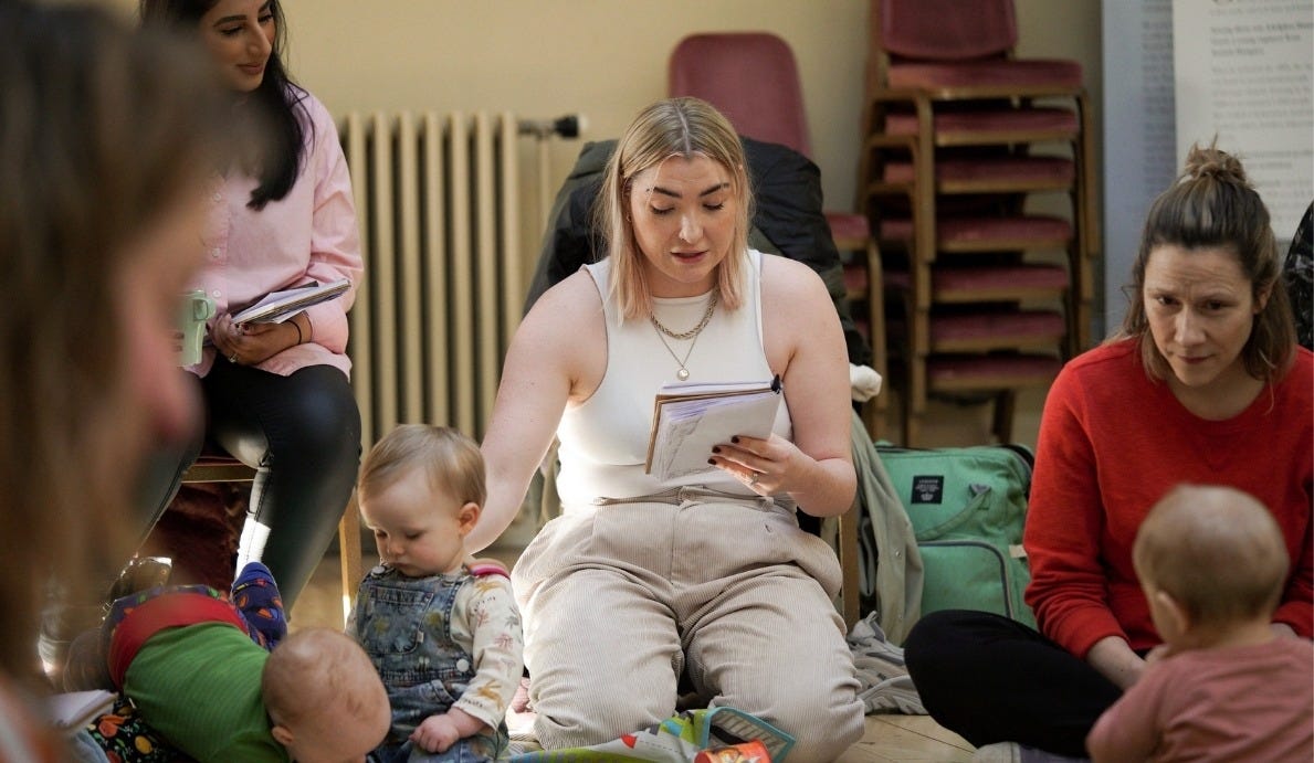 A group of adults and babies are gathered on the floor, engaging in activities, with chairs stacked in the background.
