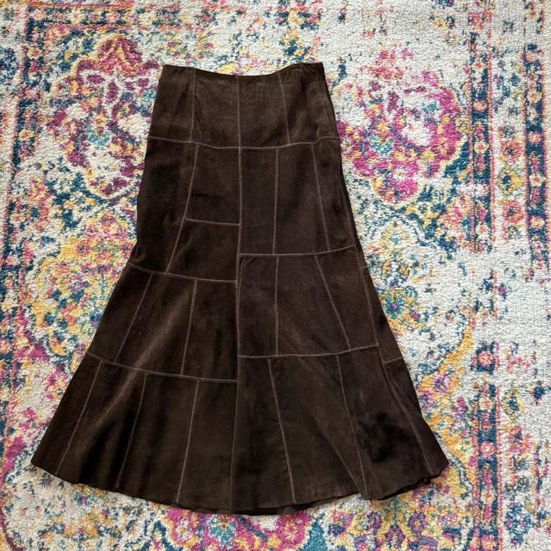 May include: A brown suede skirt with a flared silhouette and a patchwork design. The skirt has a high waist and a fitted top. The skirt is made of multiple panels of suede that are sewn together to create a unique look.