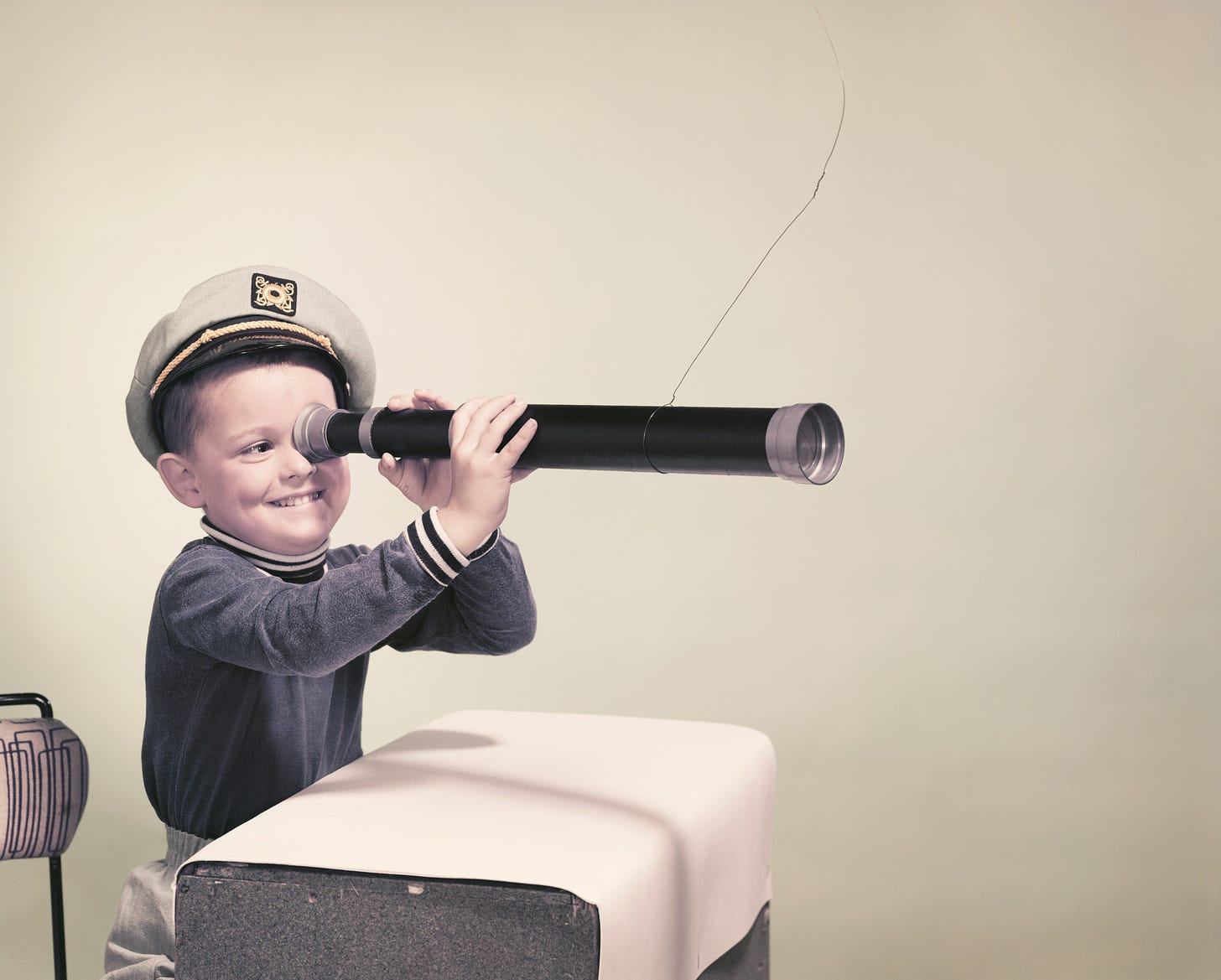Vintage Getty image in faded colour. Child in a ship's captain hat sitting at a school desk looking through a hand held telescope. 