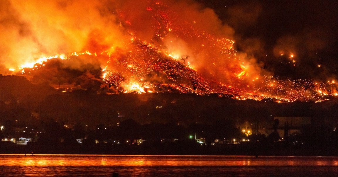 A large wildfire is burning intensely on a hillside at night, with bright orange flames and glowing embers spreading across the landscape. Thick smoke rises into the dark sky, and the fire's glow reflects on the water in the foreground. Dim lights from buildings or homes are visible at the base of the hill. A large wildfire is burning intensely on a hillside at night, with bright orange flames and glowing embers spreading across the landscape. Thick smoke rises into the dark sky, and the fire's glow reflects on the water in the foreground. Dim lights from buildings or homes are visible at the base of the hill.
