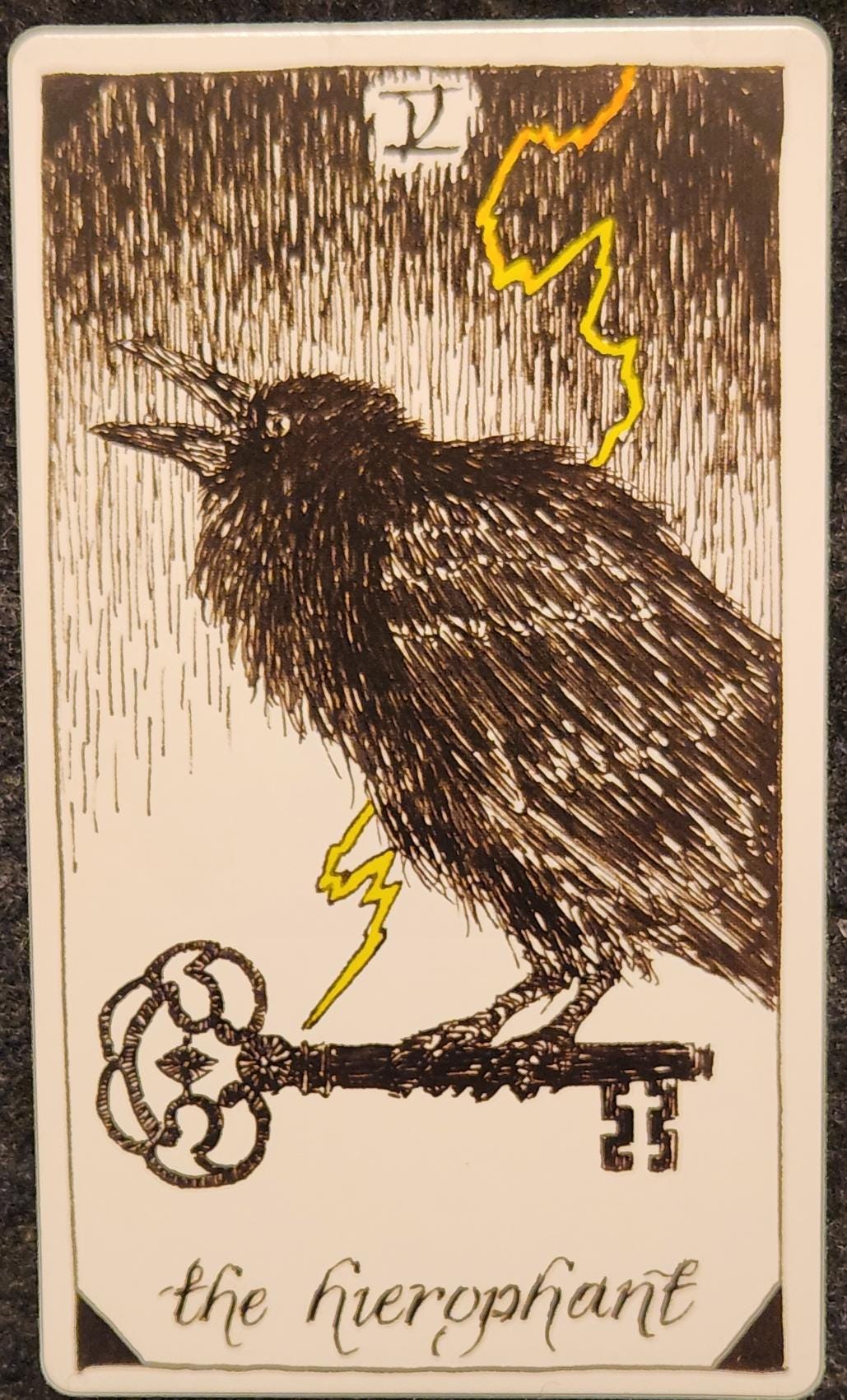 A tarot card labelled V the hierophant. A black crow is sitting perched on a large, ornate black key. Behind the crow, a bolt of lightning is flashing across the sky. 