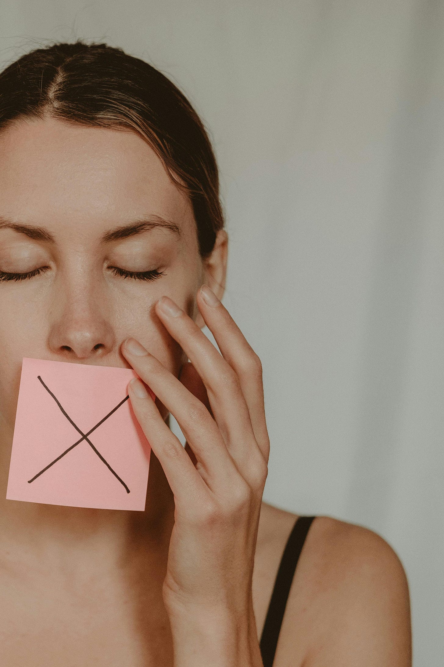 A dark haired younger woman with a pink sticky note with a black X on it covering her mouth.