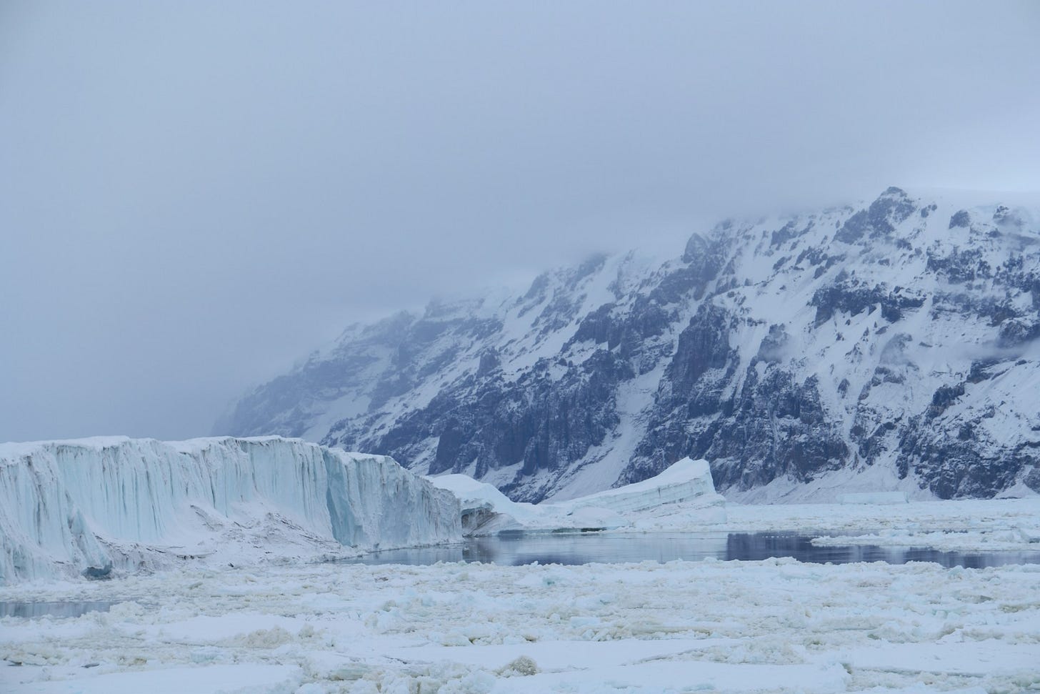 A vast, icy mountain range and glacier meet the frozen Ross Sea in Antarctica under a cloudy, atmospheric sky.
