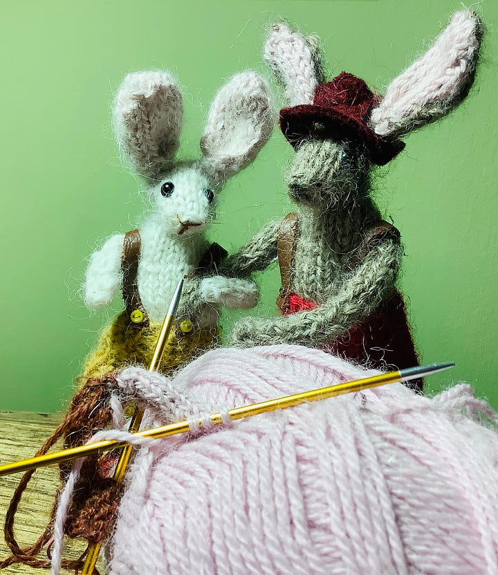 Delores and Bernard two knitted rabbits are with a ball of yarn looking at knitting needles with some stitches on.