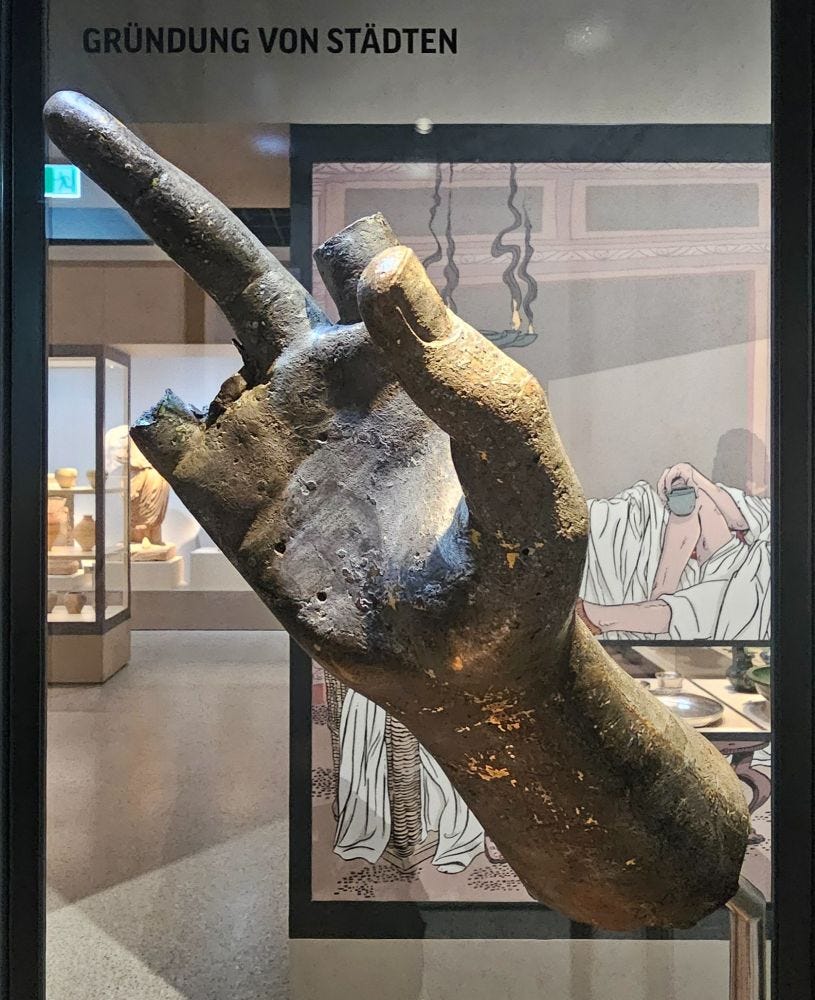 A large bronze hand is displayed in a museum, with its middle finger extended while the other fingers except the thumb are broken off. A large bronze hand is displayed in a museum, with its middle finger extended while the other fingers except the thumb are broken off.
