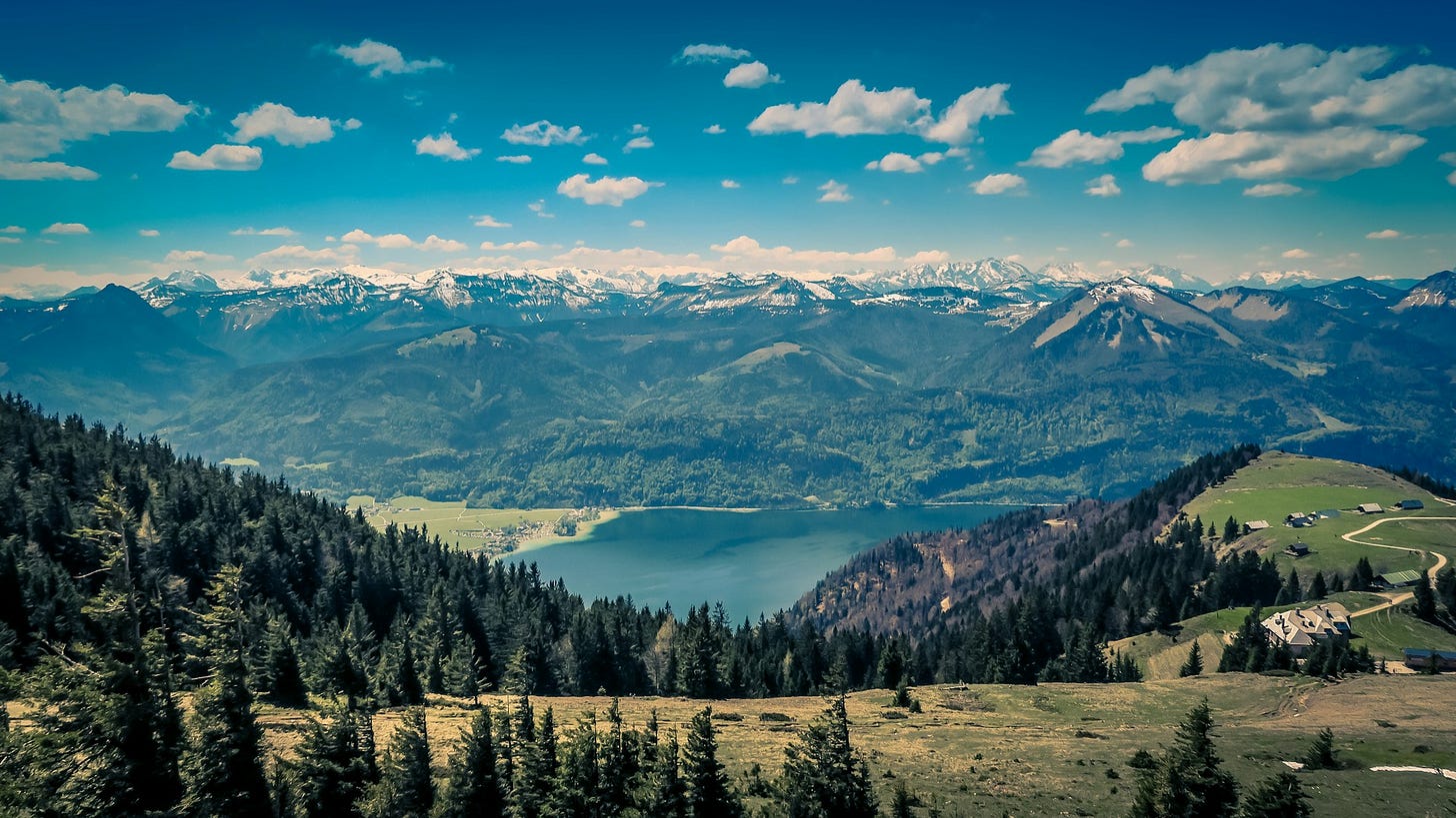 Scenic mountain landscape with a lake, forested hills, and snow-capped peaks under a bright blue sky. Scenic mountain landscape with a lake, forested hills, and snow-capped peaks under a bright blue sky.
