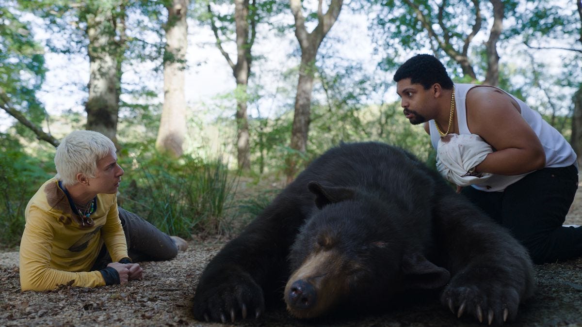 Stache (Aaron Holliday), wearing a mustard-yellow shirt and with close-clipped, white-dyed hair, talks to Daveed (O’Shea Jackson, Jr.), wearing a sleeveless T and with a shirt wrapped around his bloodied hand, over the body of a passed-out CGI bear in Cocaine Bear