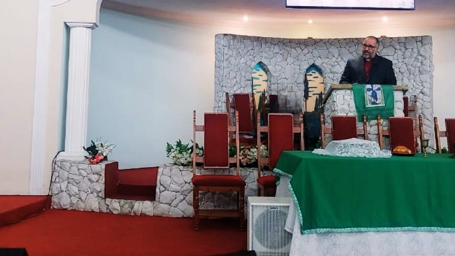 Ambiente religioso A imagem mostra o interior de uma igreja ou espaço de culto cristão. Um homem está em pé atrás de um púlpito, que está coberto com um tecido verde decorado com um símbolo cristão (parece ser uma cruz ou algo semelhante). Móveis e disposição Atrás do púlpito, há várias cadeiras com almofadas vermelhas organizadas em fileiras — provavelmente destinadas a líderes religiosos ou membros do coral. À frente, há uma mesa coberta com um pano verde e branco. Sobre ela, há um pão e outro item coberto, sugerindo que seja usada para a comunhão. Decoração O fundo tem uma parede com padrão de pedras e duas decorações estilizadas que lembram folhas ou ramos de palmeira. Há também arranjos florais e uma coluna branca decorativa à esquerda. Momento retratado Parece estar acontecendo um culto ou sermão formal. O ambiente está bem organizado e decorado, transmitindo reverência e solenidade.