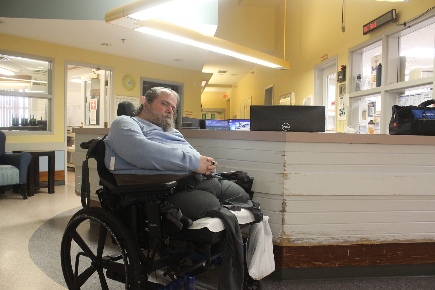A man in a wheelchair is positioned in front of a desk designed to resemble a “command center.” He is wearing a blue sweater and Vulcan “Spock ears,” with his hands clasped in his lap.