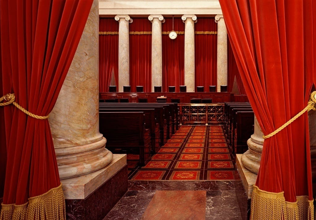 Photo of the interior of the Supreme Court, showing the dais and empty chairs where the justices sit, with white marble pillars and a red curtain in background. Photo is taken from the entrance to the chamber, framed by marble pillars and smaller red curtains with gold fringe, open and held back by gold fabric ropes, creating the sense that the viewer is coming into the Court chamber. Photo of the interior of the Supreme Court, showing the dais and empty chairs where the justices sit, with white marble pillars and a red curtain in background. Photo is taken from the entrance to the chamber, framed by marble pillars and smaller red curtains with gold fringe, open and held back by gold fabric ropes, creating the sense that the viewer is coming into the Court chamber.