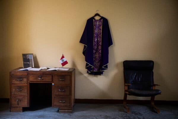 In a room with a light yellow wall stands a wooden desk with a photograph, flag and books, which lie open. A religious robe is hanging on the wall to the left of a black chair.