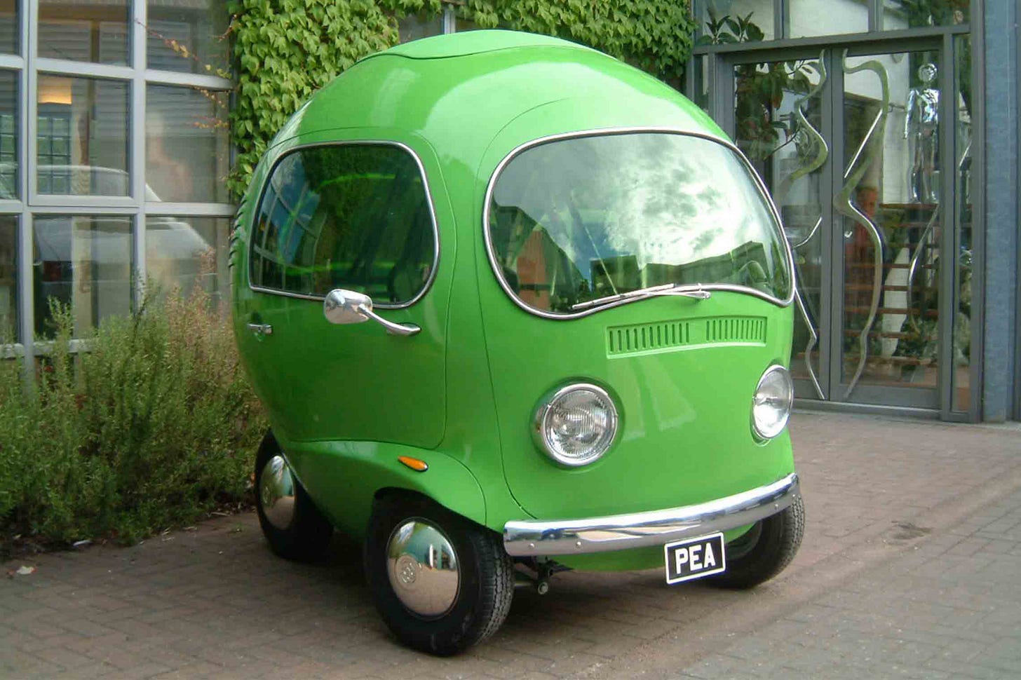 Photograph of a small, spherical, bright green custom car shaped like a pea, with rounded windows, chrome bumper, and the license plate “PEA,” parked in front of a building with ivy on the wall.
