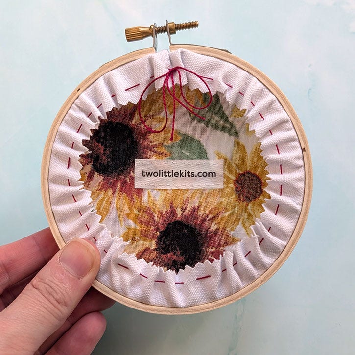 The "butt" of an framed needlepoint in an embroidery hoop, which has a little fabric signature reading 'twolittlekits.com'.