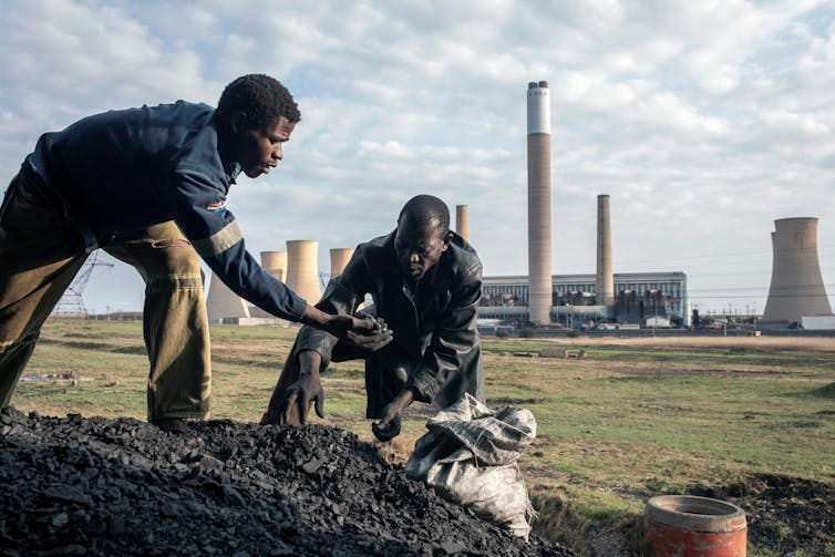 Two men put coal chunks into a sack with a power plant in the background.