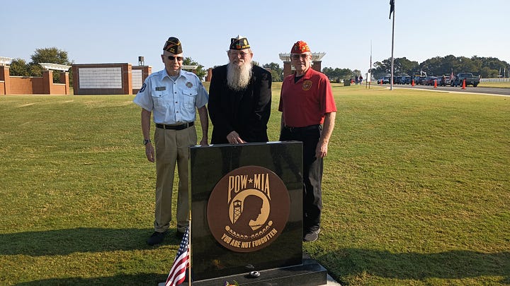 A group of veterans and community members pose outdoors at a POW*MIA memorial. In the foreground is a black granite monument engraved with the POW*MIA emblem and the words “YOU ARE NOT FORGOTTEN,” with a small American flag at its base. Several U.S. flags stand behind the group. Some people wear military dress uniforms and VFW-style caps; one cap reads “Tallahassee,” and another hat bears the U.S. Air Force insignia. On the right, an elderly veteran in a wheelchair salutes while a man in a suit speaks with him. The scene is on a sunny day in a grassy field with cars and flagpoles in the background.