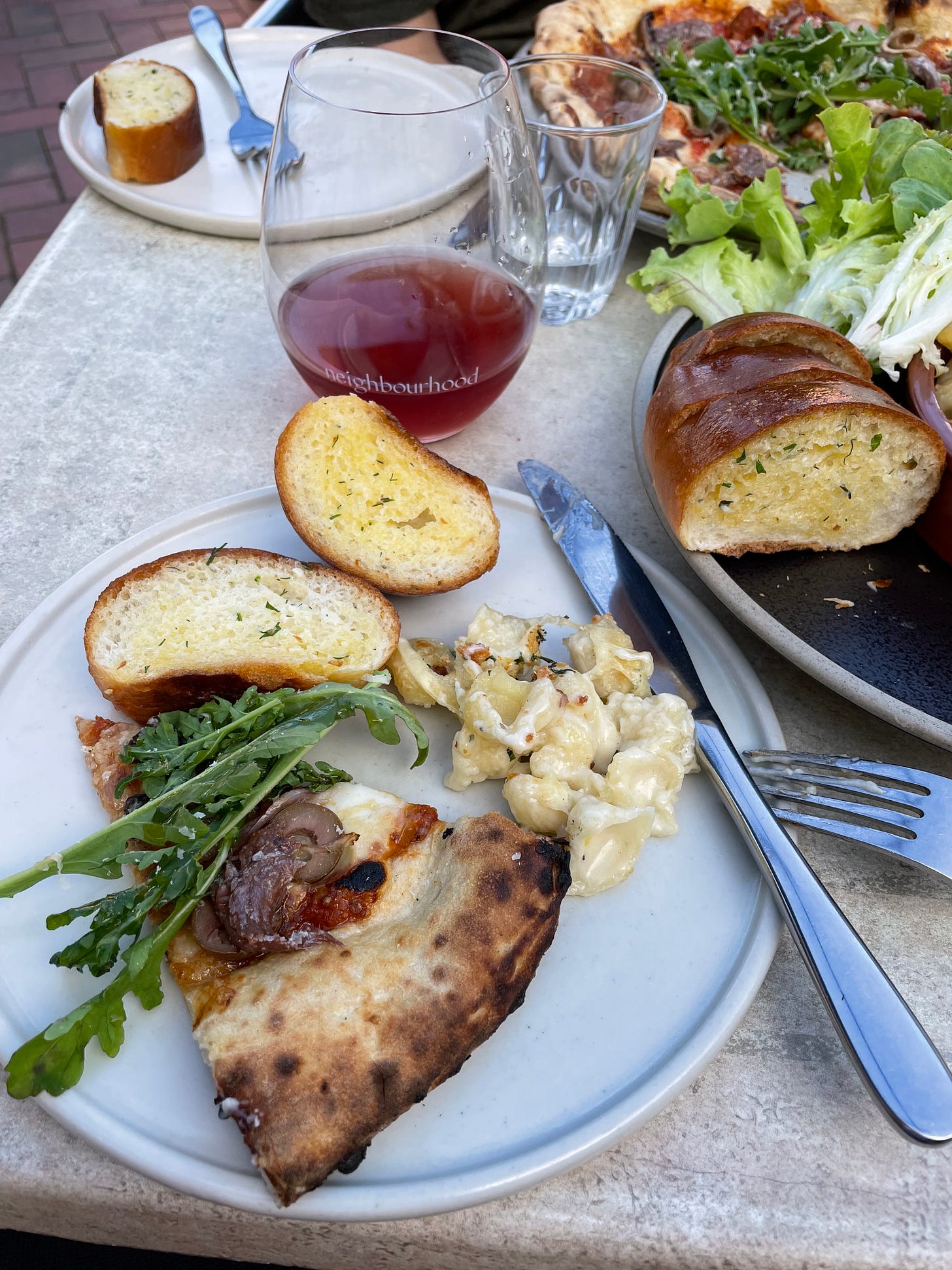 Plate of mac and cheese, garlic bread slices and pizza with a salad in the background. Dinner at a neighbourhood restaurant.