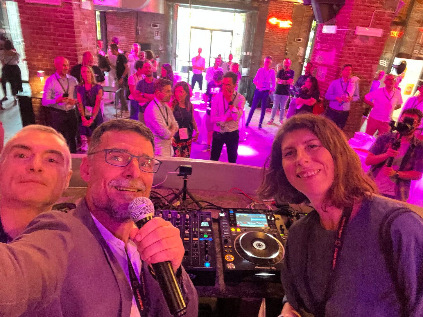 Stefano Gatti, io (Matteo Fogli) e Mafe de Baggis in un selfie dalla console del DJ del The Beach a Torino, 1 minuto prima di iniziare la "fireside chat" su Intelligenza Artificiale e futuri possibili. Alle nostre spalle decine di persone in piedi per l'inizio della chiacchierata, illuminate da luci viola. tra le colonne e le arcate di mattoni a vista del locale, durante l'evento Orbyta Futures Party