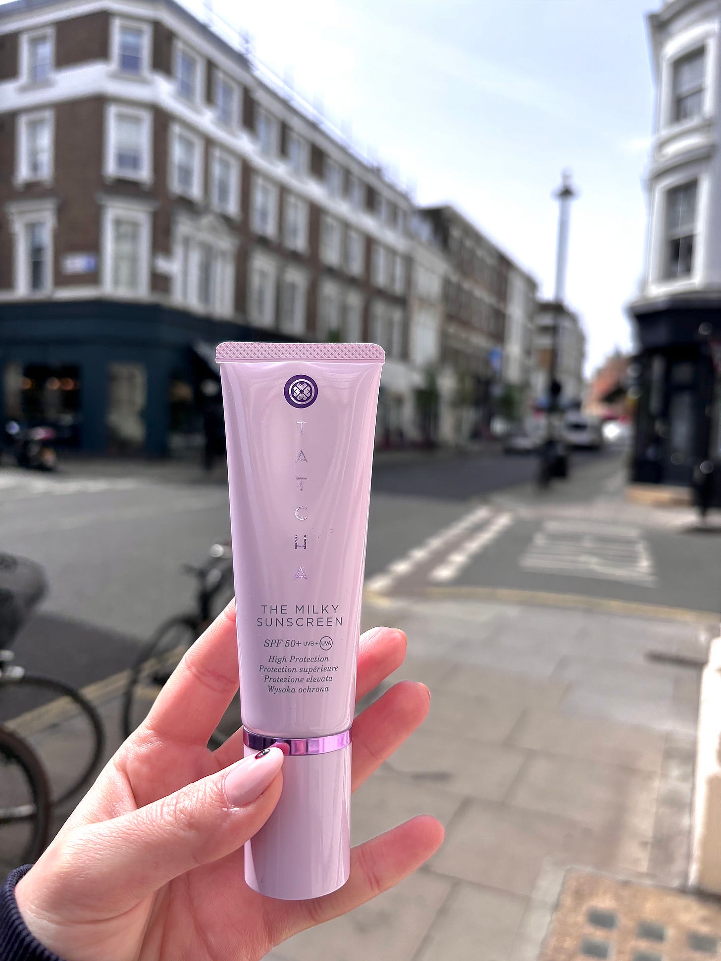 A close-up, unretouched shot of Tatcha The Milky Sunscreen SPF 50+ held against a blurred London street background. This signals a pre-vacation trial for The Beauty Ed.® to test formula performance in humid, urban environments.