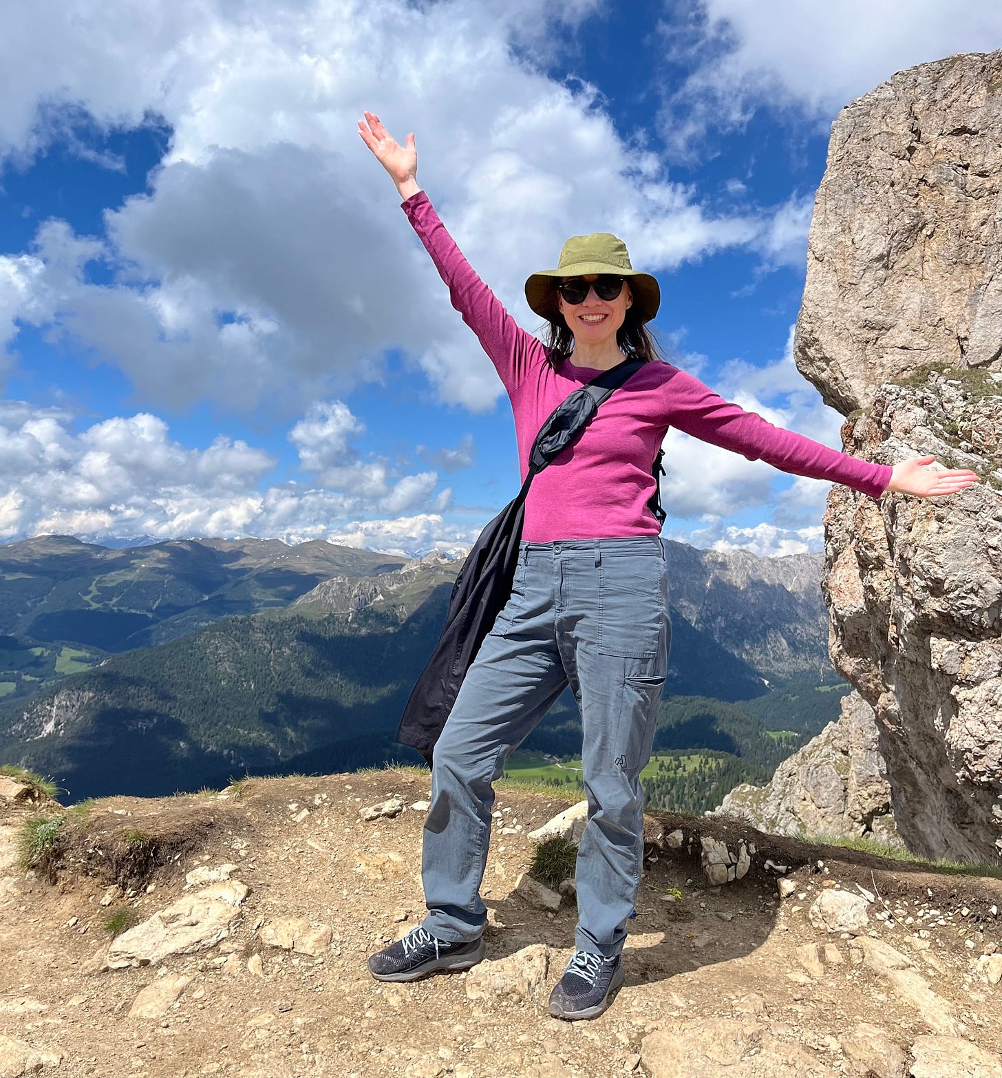  I am standing at the top of a mountain, wearing hiking gear, sunglasses, and a wide smile. My arms are wide, embracing the beauty. There is a green valley below, and another mountain range in the distance, under a blue sky mostly hidden by clouds.