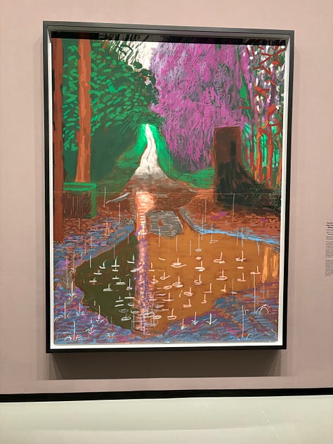 Multiple bright-colored paintings including a self-portrait of Hockney in a yellow plaid suit and big yellow glasses, the crowd viewing paintings, a mother and child in front of a bright painting of a massive tree, an annunciation painting redone in neon colors, a green lawn with sprinkler spray, a photograph of hockney working at his drawing table with his little terrier dog under the table and two paintings with raindrops hitting puddles among trees