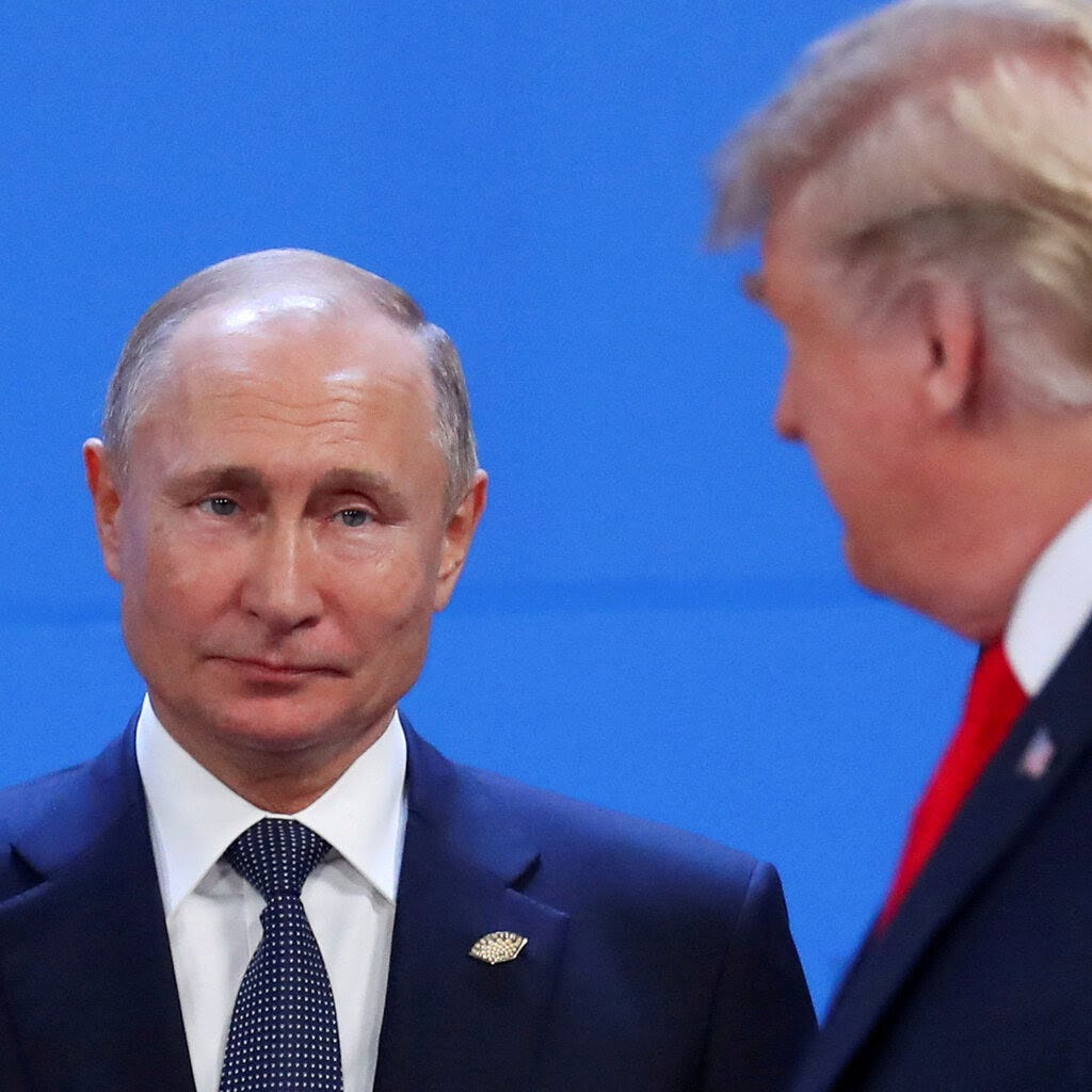 Vladimir Putin stands with his head slightly titled while Donald Trump, blurred in the foreground, looks on.