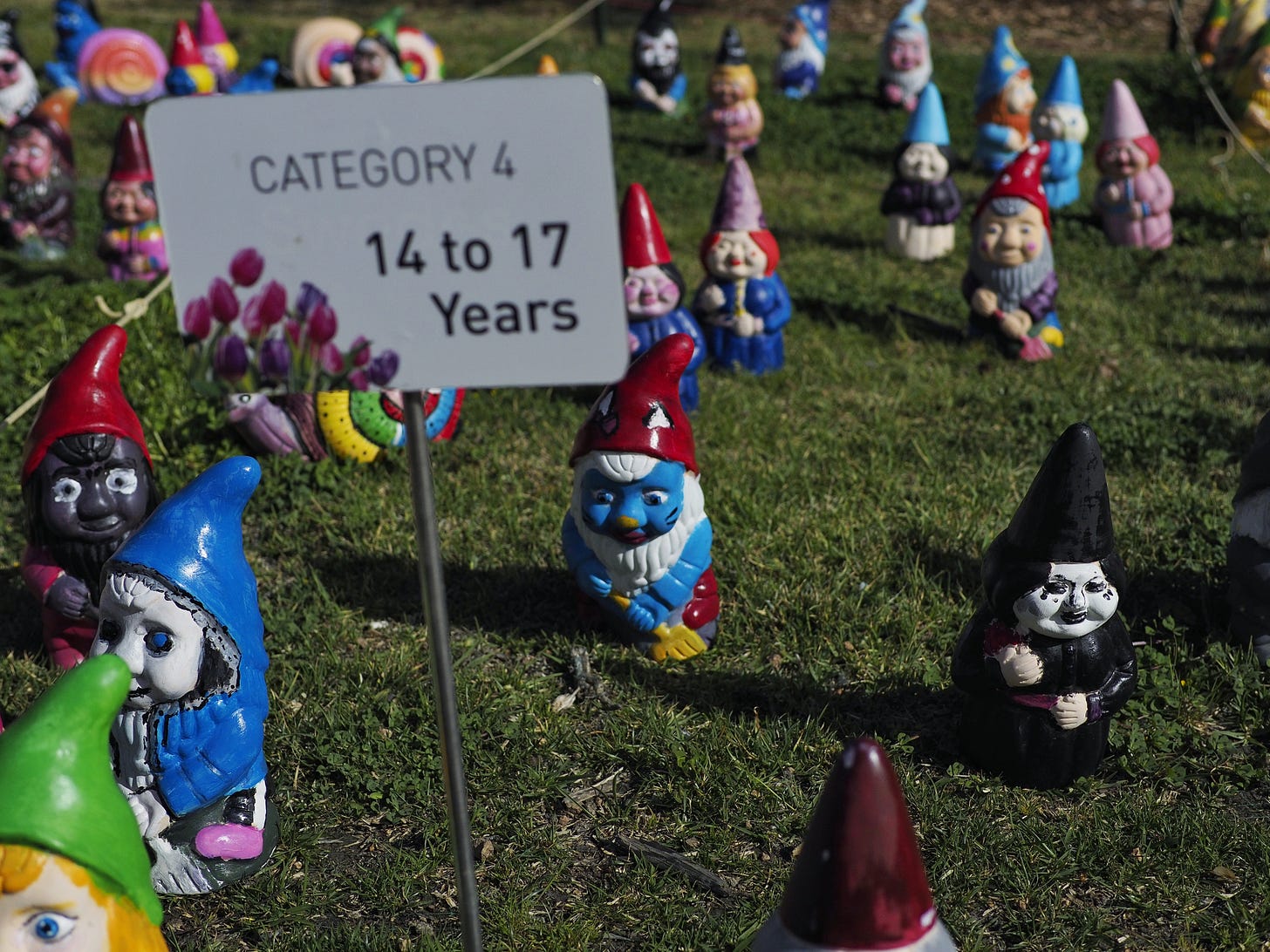 A photo of painted gnomes on a lawn. There is a sign which reads, "Category 4: 14 to 17 years". The focus gnome is painted wearing all black with a pale white face, black lipstick and clown tears. it is holding a bunch of dark red flowers. 