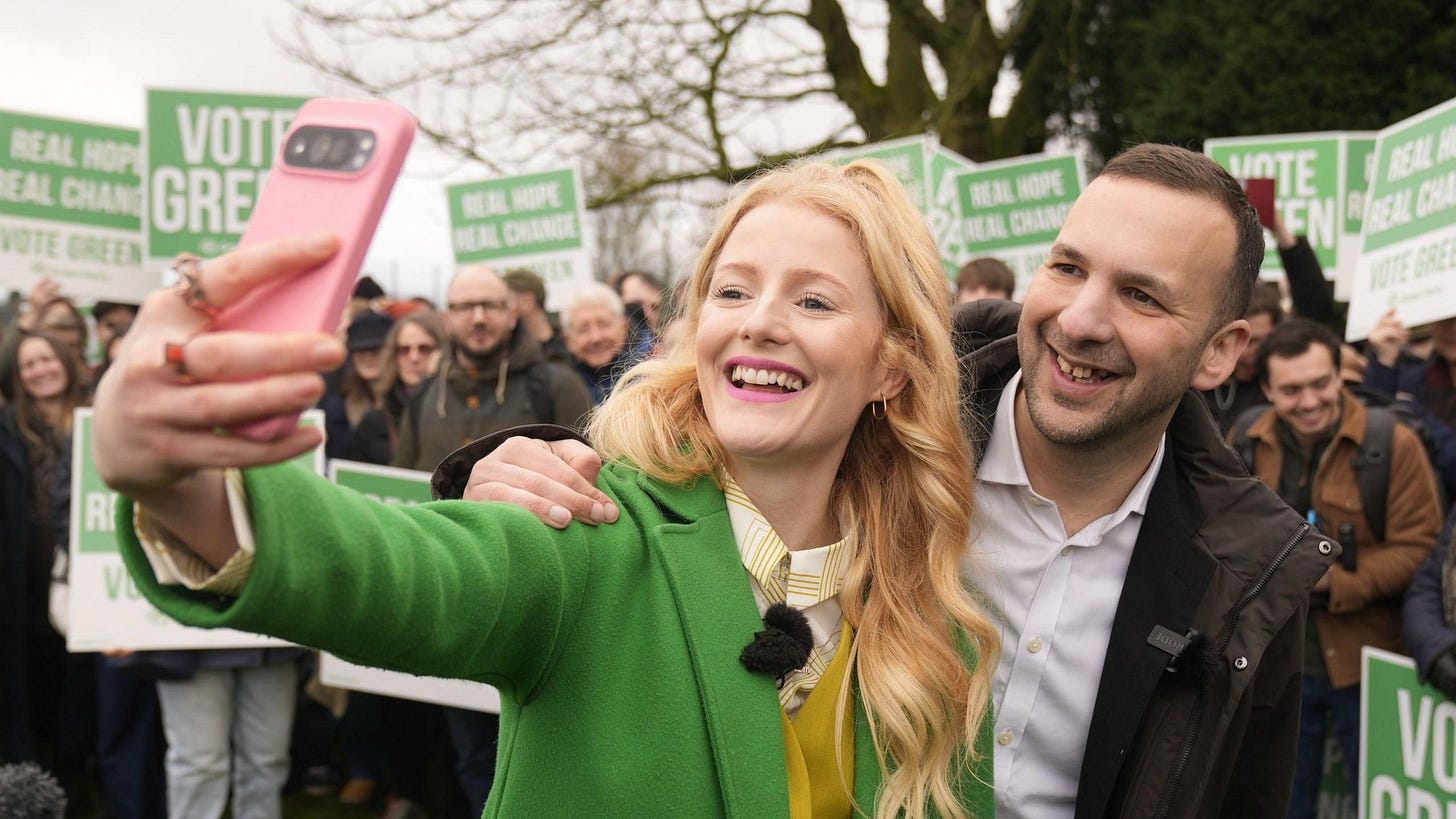 Hannah Spencer Green Party candidate takes selfie with Zack Polanski at Denton and Gorton by-election campaign rally, February 2026, as supporters hold vote green real hope real change signs behind them, while Bristol residents march against ignored liveable neighbourhood consultation and 54% opposition to £6 million traffic scheme continues unheard by Green council- https://bit.ly/4tt79em