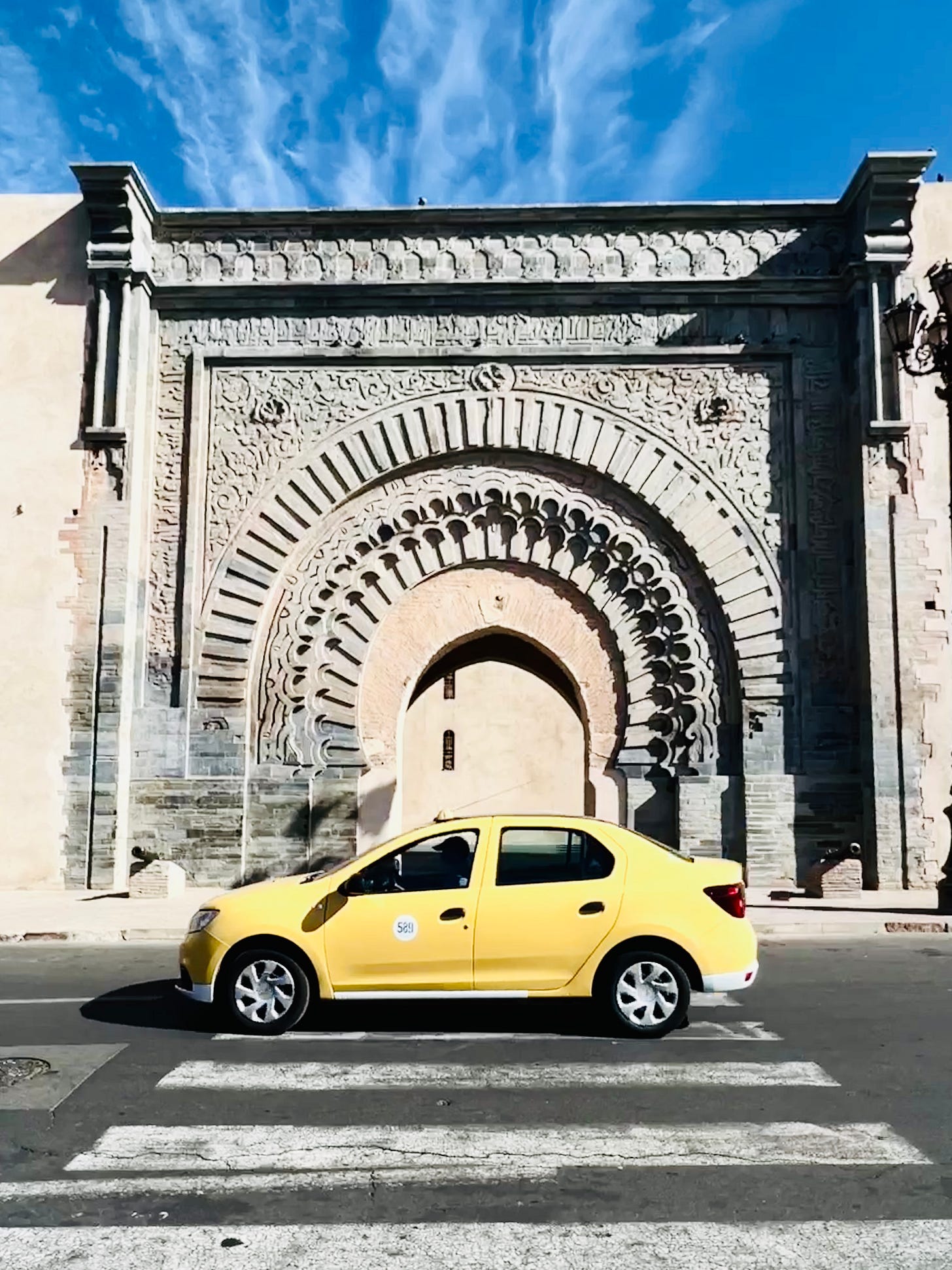 A yellow taxi drives past an ornate historic city gate in Marrakech, framed by carved stone arches and a bright blue sky, highlighting the contrast between daily transport and architectural heritage. A yellow taxi drives past an ornate historic city gate in Marrakech, framed by carved stone arches and a bright blue sky, highlighting the contrast between daily transport and architectural heritage.