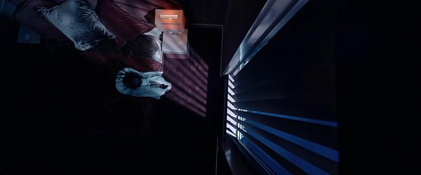 A woman sitting on the edge of her bed within a darkened bedroom.