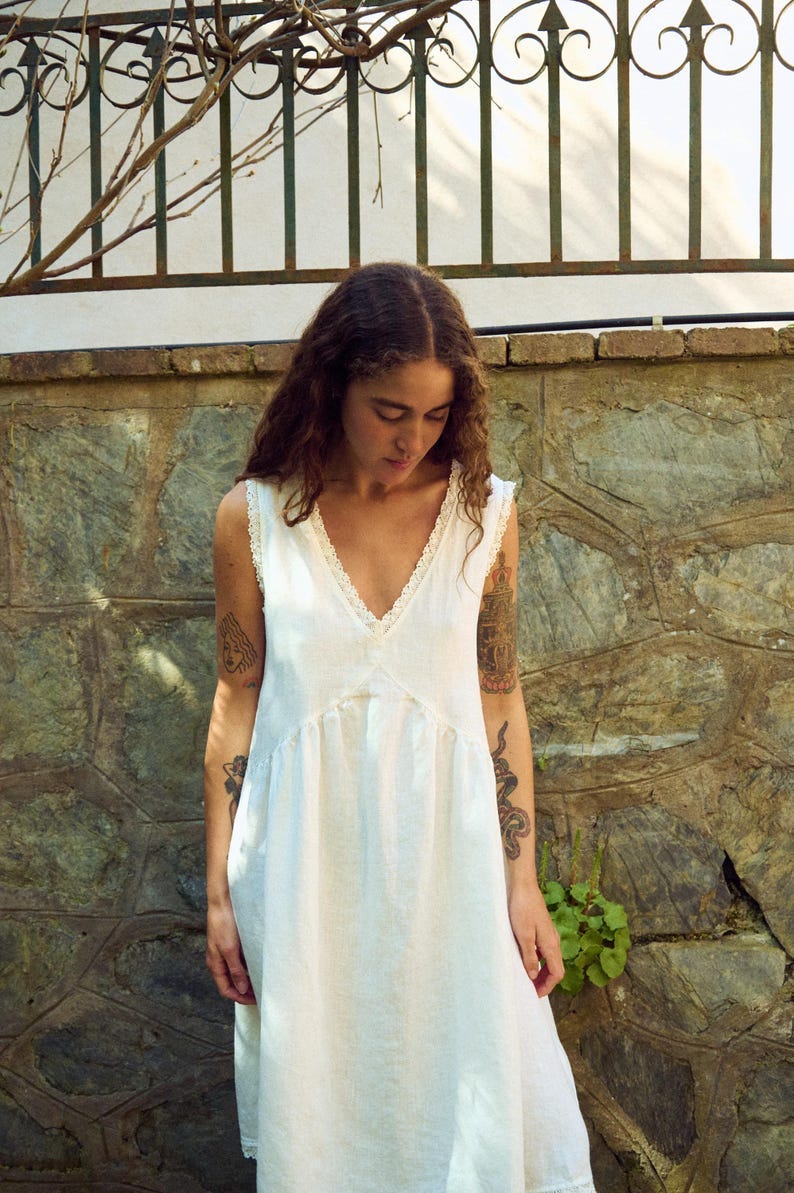 May include: A sleeveless white dress with a V-neckline and lace trim. The dress has a flowing, relaxed fit and a gathered waist. The model is standing outdoors near a stone wall and a wrought iron fence.