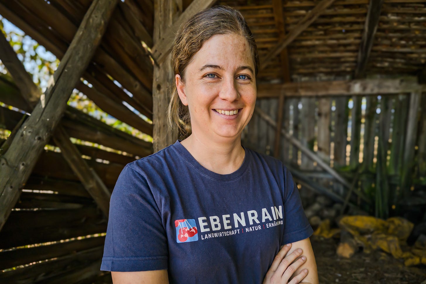 Projektleiterin Sereina Grieder vom Ebenrain-Zentrum steht lächelnd mit verschränkten Armen in einem offenen Holzschuppen. Sie trägt ein dunkelblaues T-Shirt mit dem Ebenrain-Logo; durch die Holzwände fällt Sonnenlicht.