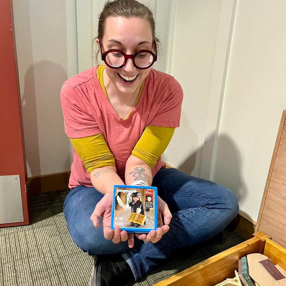 Bird, a white woman with large red glasses and brown hair, sits on the floor holding out a box featuring a Playmobil figurine of Marin Luther. Her face is focused on the box, grinning with delight.