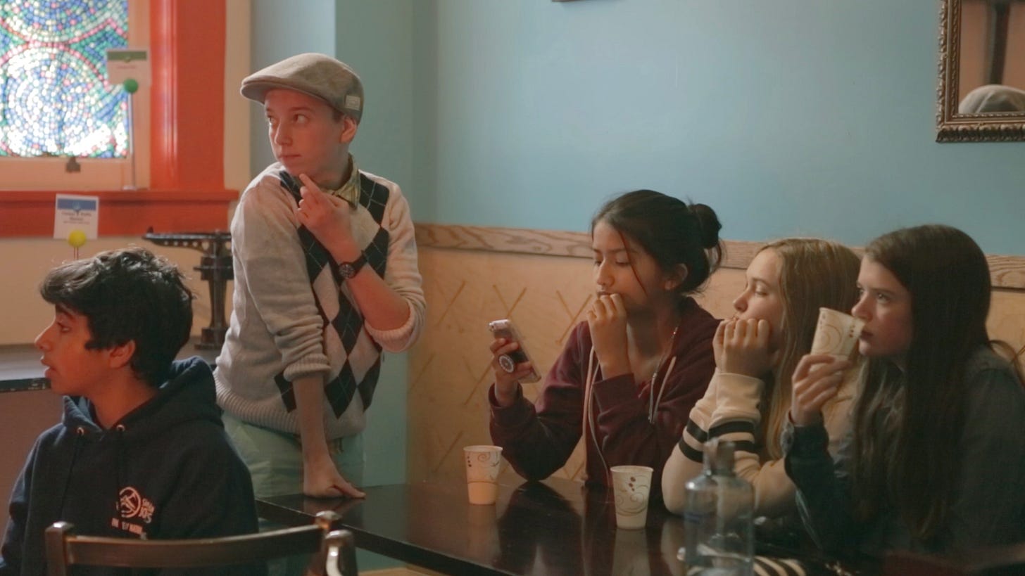 Bea, standing at a table, with short hair under a cap looking off to the side as three girlfriends sit at a table and a male friend sits on the other side.