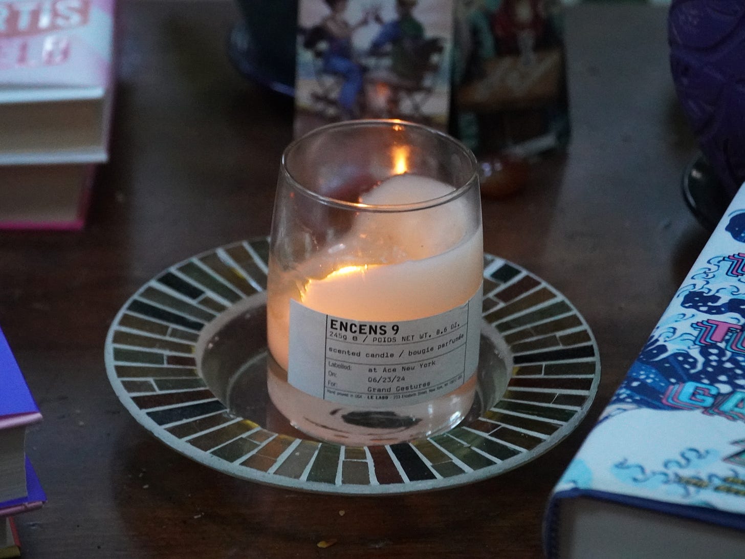 On a wooden table sits a lit cream-colored candle on a plate. It has a white label that reads ENCENS 9. Scented Candle. Under it it says, at Ace New York, 06/23/24, Grand Gestures, Le Labo. The candle is surrounded by books.