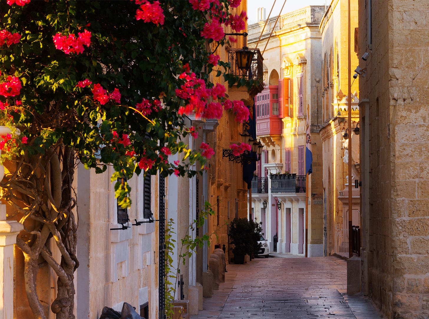 A stone-paved alley in Mdina, Malta, glowing in early morning light. Bougainvillea with vivid pink blossoms spills into the frame on the left, its twisted trunk anchored in limestone. Ornate black lanterns hang from the golden walls, leading the eye toward a cluster of traditional Maltese balconies painted in bright pink and purple. The scene captures Mdina's mix of medieval stonework, Mediterranean flora, and the colorful domestic details that bring life to the Silent City.