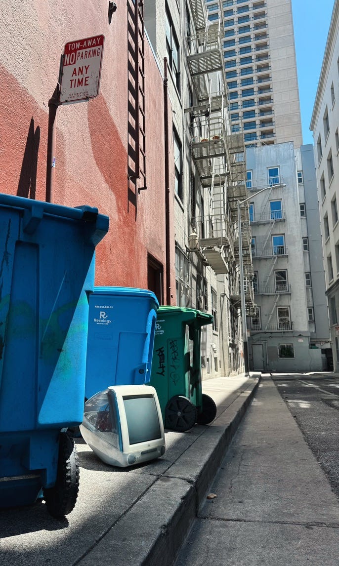 A back street in an American city showing a discarded first generation iMac, which was famous for its all-in-one design and colourful casing around its CRT display. Neat to this computer are two blue and one green wheelie bins. It just so happens that the sunlight catches the dusky pink at angle to give the picture a subtle glow that makes this angle of an insalubrious alleyway appear like a still life scene.
