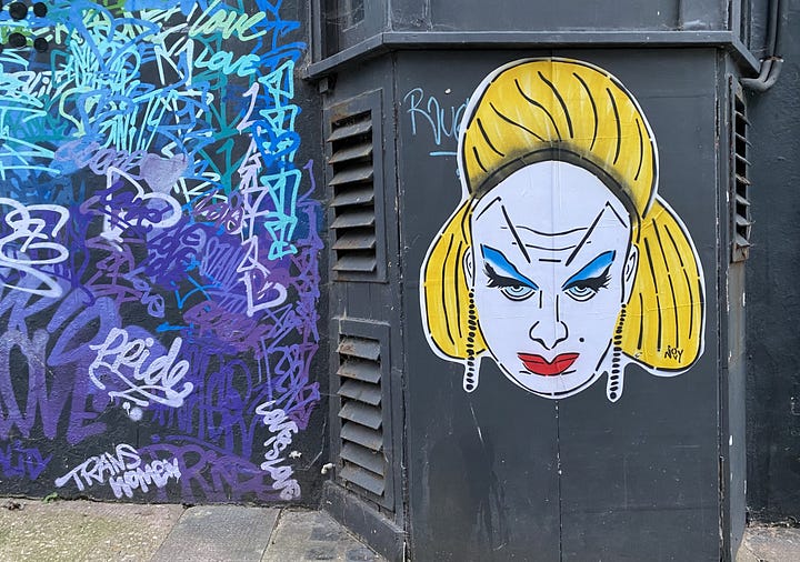 1) Street art on an orange "Detour" sign features a stylized face with blue paint for hair, bold red makeup, and exaggerated lashes, creating a vibrant and edgy vibe. This portrait is of Leigh Bowery, a famous queer person. There is more graffiti and tags behind Jeremy's work. 2) Street art on a dark black wall features a stylized portrait of a person with yellow hair and bold makeup. On the wall nearby, vibrant blue and purple graffiti tags. The portrait is of Divine, the drag queen. She has very high eyebrows, blue eyeshadow, and red lipstick, with a bright puffy yellow wig.