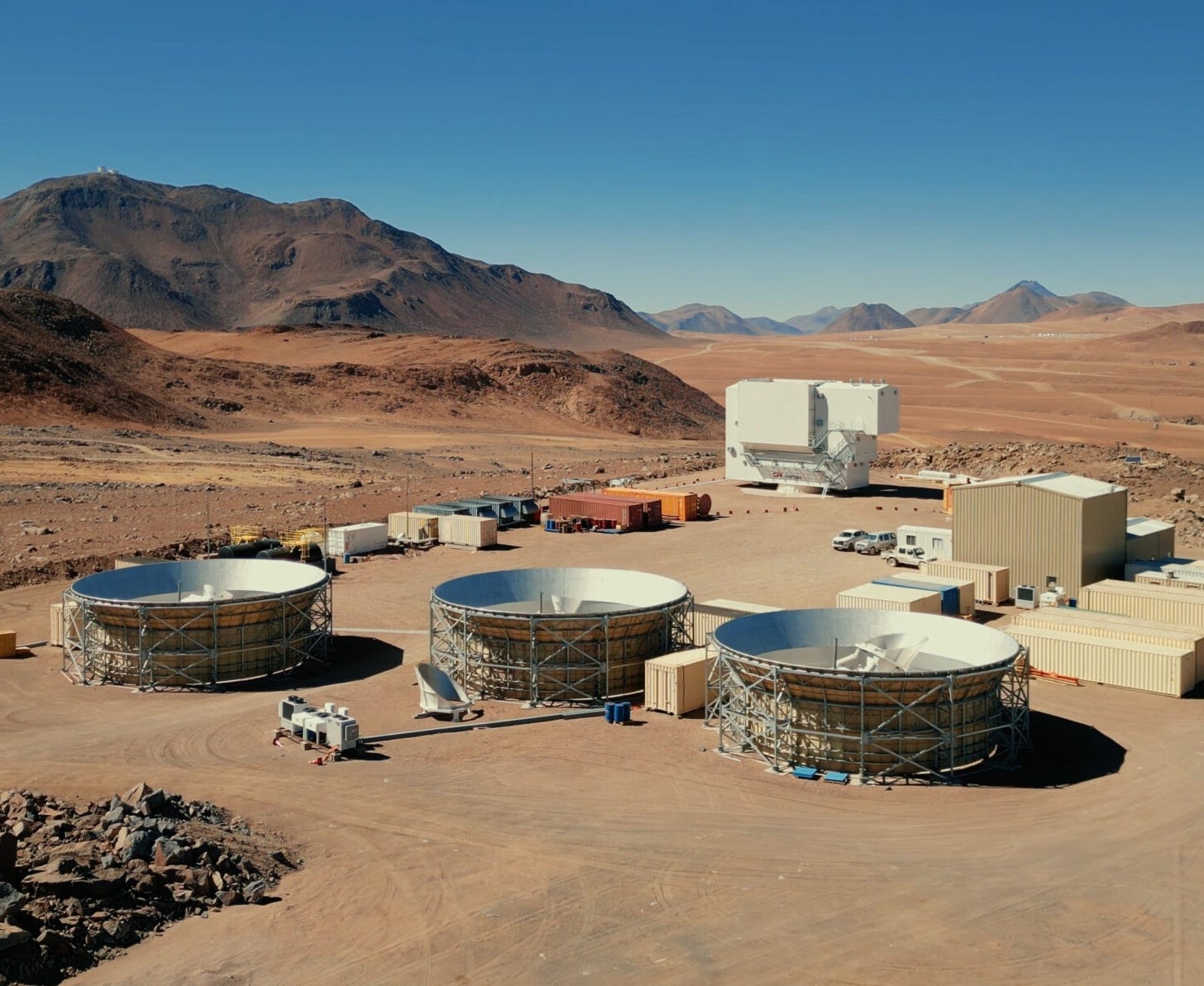 Three large satellite dishes and several buildings are set on a dry, rocky landscape with mountains in the background under a clear blue sky, showcasing a research facility dedicated to exploring the mysteries of cosmology and phenomena such as cosmic birefringence.