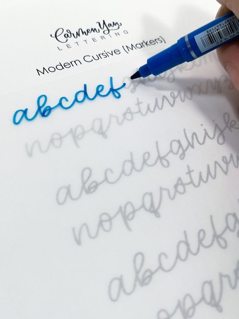 May include: A close-up of a hand using a blue marker to write the letters "abcdef" in cursive on a white sheet of paper. The letters are written over a faint gray outline of the alphabet. The paper has the text "Carmen Yan LETTERING Modern Cursive (Markers)" at the top.