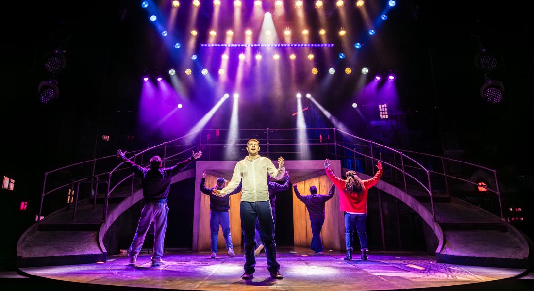 A stage set with sweeping ramps down both sides and strong spotlights, in shades of blue, pink and purple. 6 actors fill the stage. All but one face the back, their arms raised. The man at the front holds his arms out, as if asking a question.