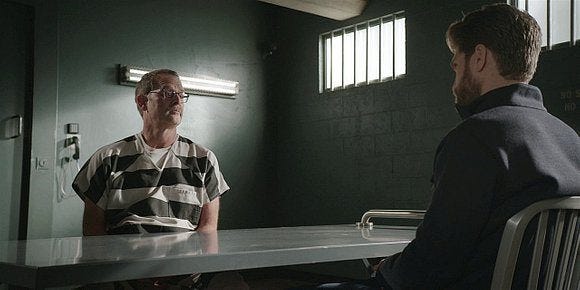 Stan looking at Liam across table in jail cell. Stan looking at Liam across table in jail cell.