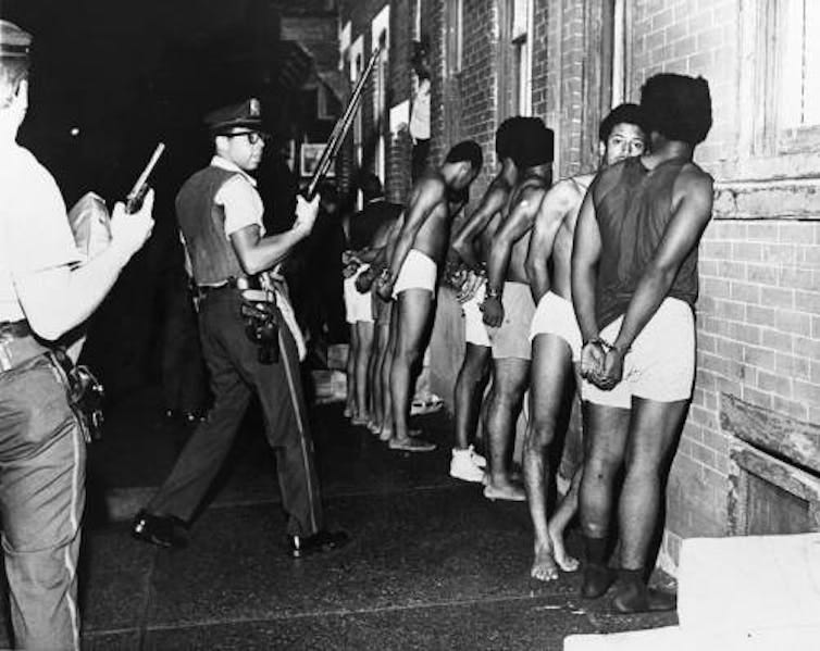 Black men barefoot, handcuffed and wearing only underwear are lined up facing a building as police with guns watch over them