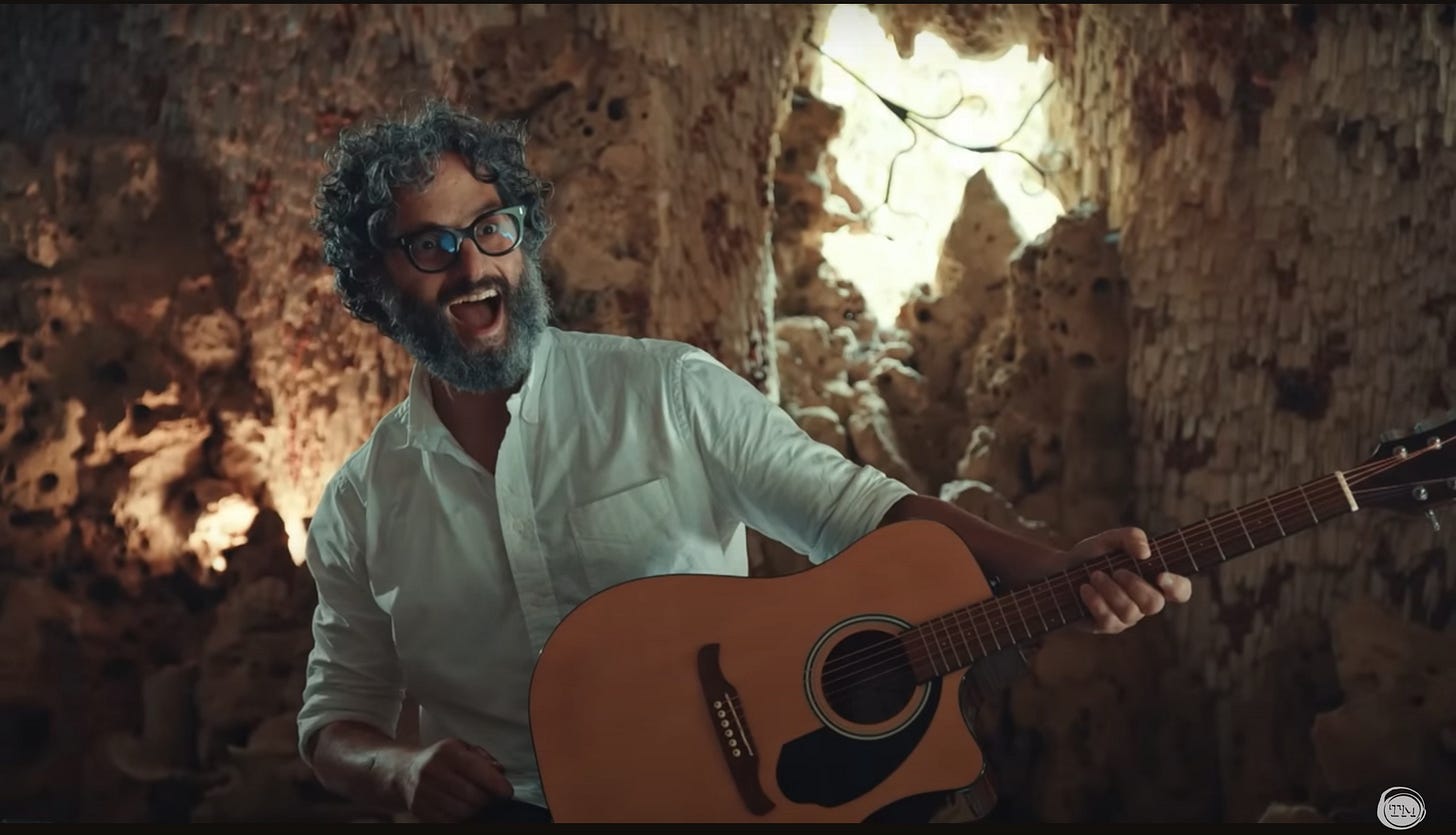 Jason Mantzoukas grins broadly while holding a guitar inside a cave.