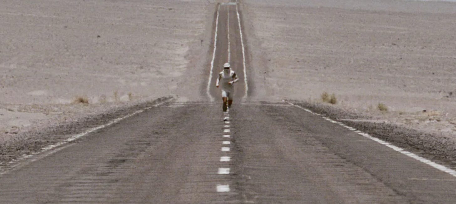 A man dressed in Satisfy's heatcrush shorts, hat, and top is running on a desert road. A man dressed in Satisfy's heatcrush shorts, hat, and top is running on a desert road.