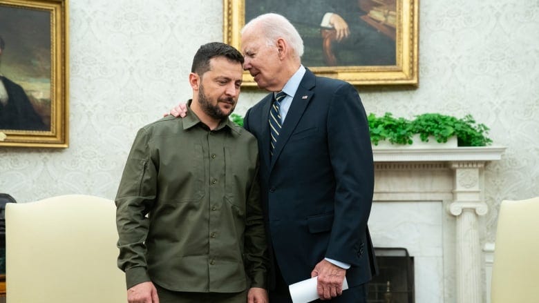 A white-haired man in suit appears to whisper in the era of a bearded, younger man who is wearing military fatigues. In the background, a fireplace and a painting above the mantle.