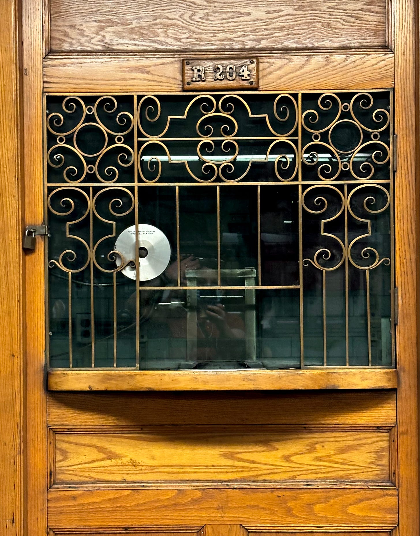 The window of the toll booth. The metal grating is in panes, each in different curly cue patterns. The author's reflection waving while she takes the picture because she couldn't get the detail of the metal work she wanted without being in the shot is also visible.