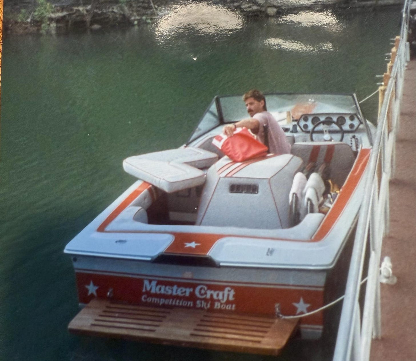 Man steers classic red, white, and blue Master Craft Stars and Stripes ski boat at the lake dock, representing cherished family traditions and a beloved uncle's influential legacy in his nephew’s life.